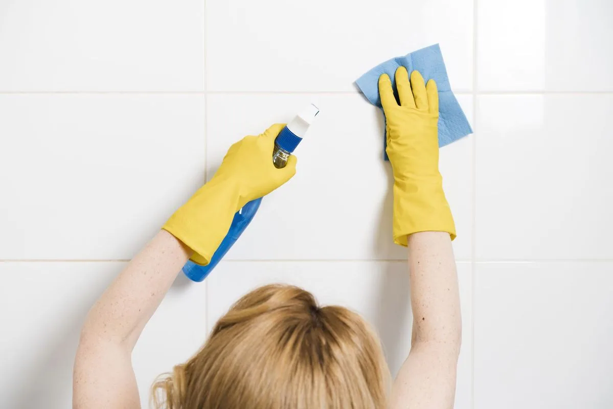 Person wearing yellow gloves sprays and wipes white tile wall with a blue spray bottle and cloth.