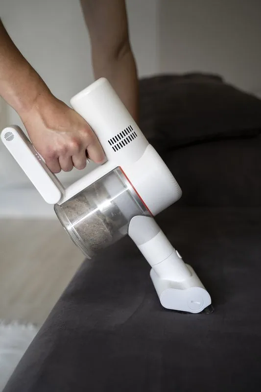 Person uses a white handheld vacuum to clean a dark gray upholstered surface.
