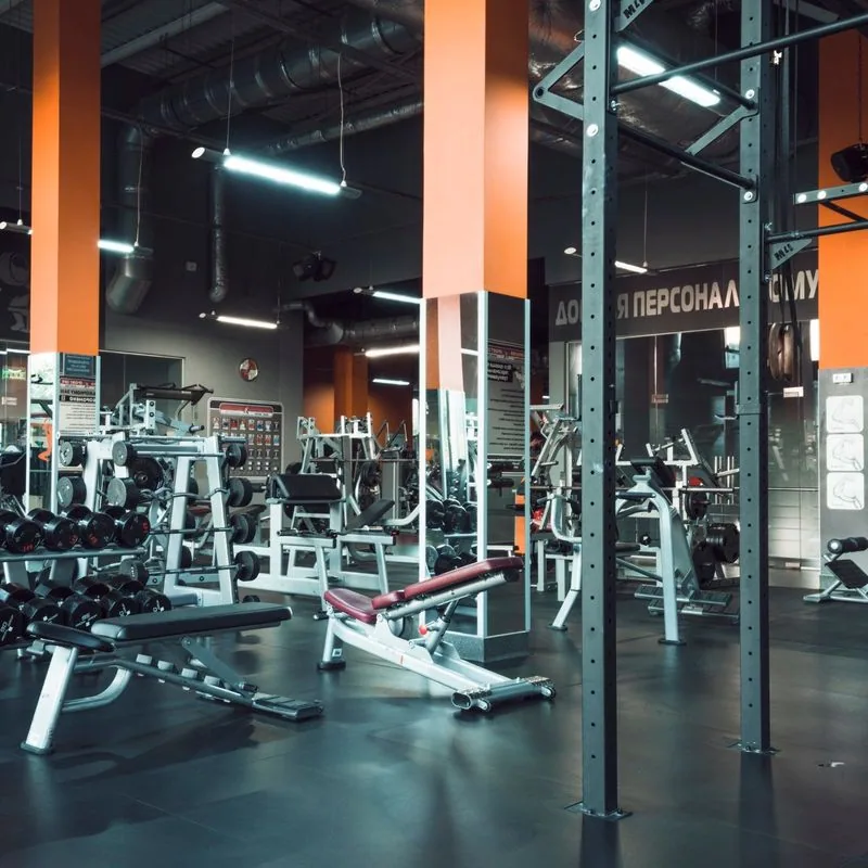 Gym interior with various weight machines, benches, and dumbbells. Orange columns and dark flooring.
