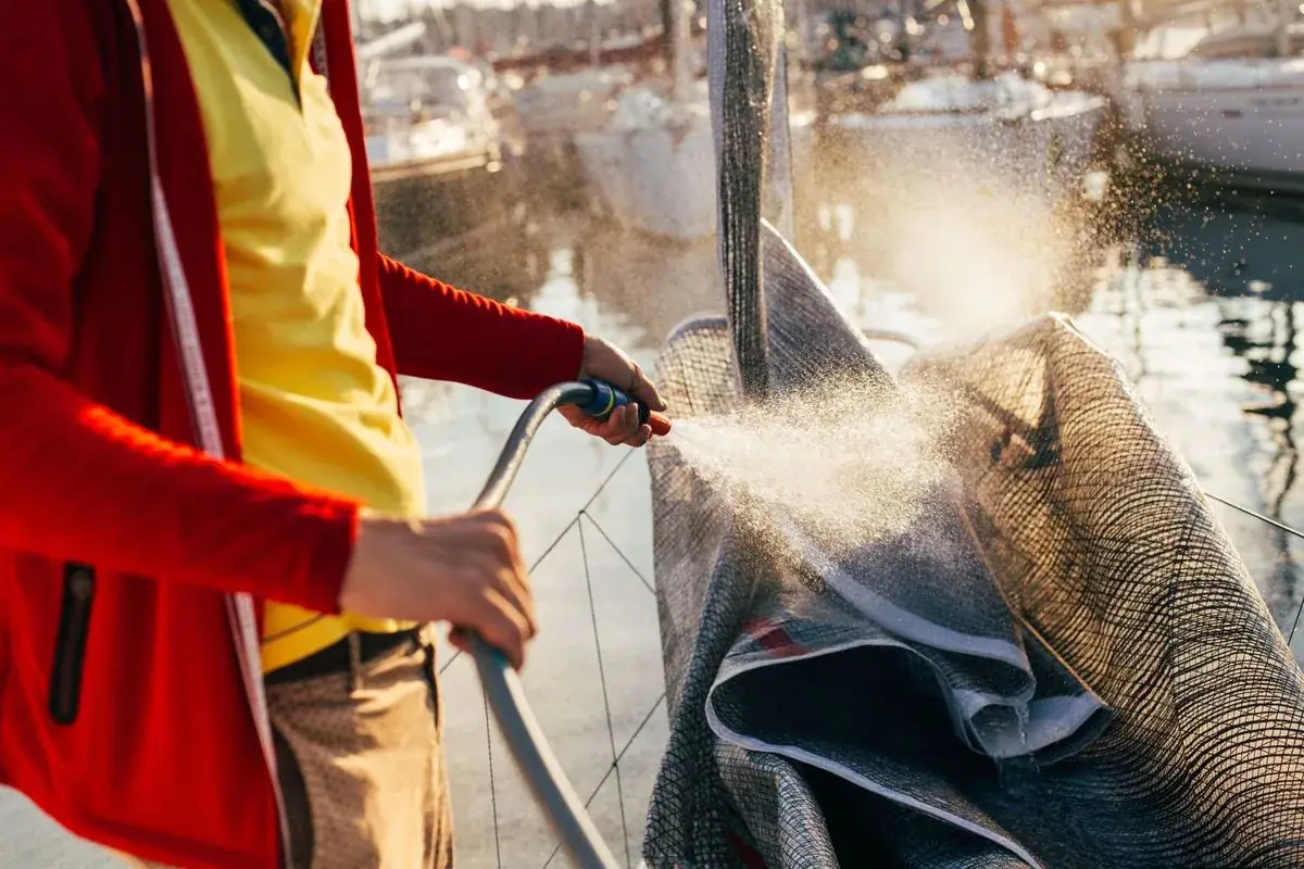 Person in red jacket sprays water on a boat sail in a harbor.