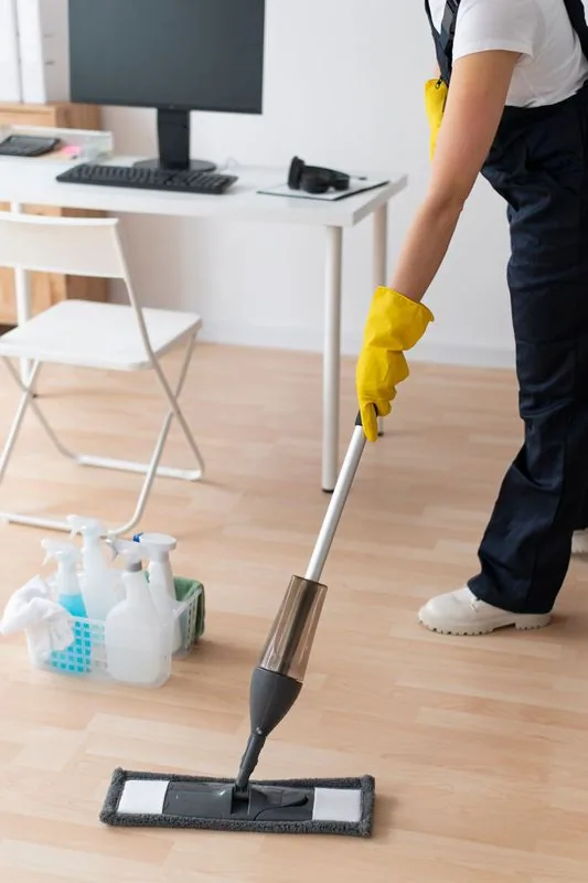 Person mopping a wooden floor in an office setting. Yellow gloves, spray mop, cleaning supplies.