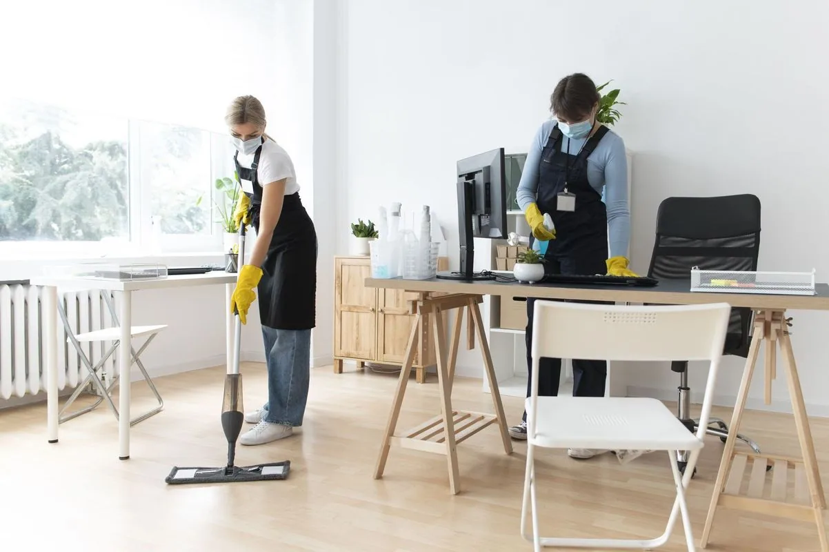 Two people wearing masks and aprons cleaning an office with a mop and wiping a desk.