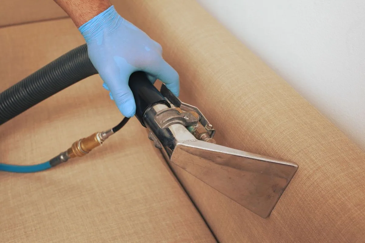 Person in blue glove vacuums a beige couch cushion with an industrial upholstery cleaner.