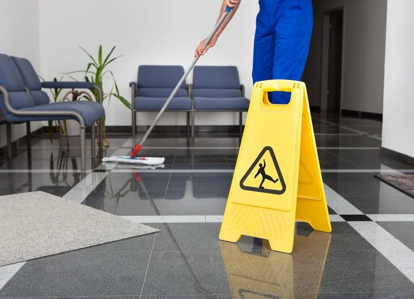 Person mopping a wet floor in an office lobby with a yellow caution sign.